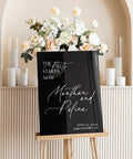 Black acrylic sign with "The Party Starts Now" and names "Manthan and Pefina," displayed in front of flowers, on an easel.