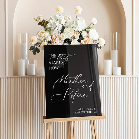 Black acrylic sign with "The Party Starts Now" and names "Manthan and Pefina," displayed in front of flowers, on an easel.