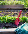 Pink shovel from Mini Colorful Metal Hand Shovels Set 10 Pack Garden Trowels in soil, beside glove, in garden bed.