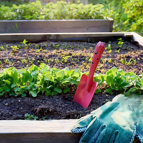 Pink shovel from Mini Colorful Metal Hand Shovels Set 10 Pack Garden Trowels in soil, beside glove, in garden bed.