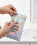 Person holding a 100 Pack Holographic Jewelry Bags With Clear Window Resealable Self Sealing Packaging For Jewelry Candy Snacks And Small Biz Gifts Iridescent Shiny Bags For Easy Identification And Stunning Display