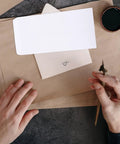 Person holding calligraphy pen near Thick White Blank Letter Envelopes 230x120mm Bulk Pack of 100 Durable Paper for Cash Envelopes Office Bill Storage and Mailing Supplies Easy Seal Clean Look for Everyday Mail on brown paper.