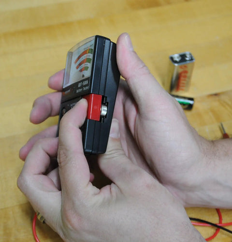 Hands operating an Analog Battery Tester With Test Leads For All Common Batteries From 1.5V Button Cells To 12V Lantern Batteries, AA And AAA, 9V, 14 Battery Range, Easy Read Meter, Black on a wooden surface.
