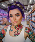 A person with purple hair and floral tattoos poses in a body jewelry warehouse, showcasing colorful ear gauges.