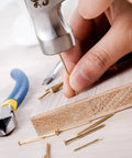 Hand using hammer with Gold Finish Nail Kit With 600 Nails In Four Sizes For Picture Hanging And Woodworking Includes Finishing Nails.