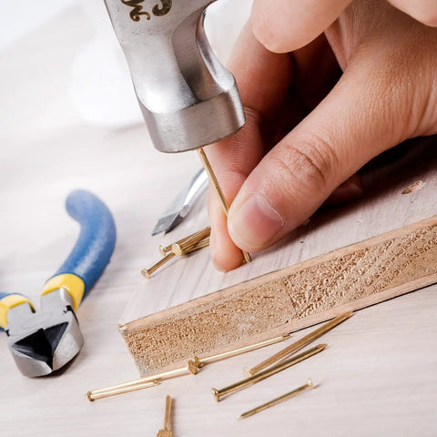 Hand using hammer with Gold Finish Nail Kit With 600 Nails In Four Sizes For Picture Hanging And Woodworking Includes Finishing Nails.