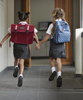 Two children wearing school uniforms and backpacks walking in a hallway holding hands.