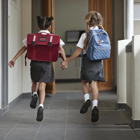 Two children wearing school uniforms and backpacks walking in a hallway holding hands.
