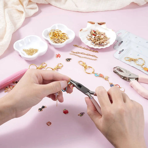 Hands using pliers with All In One Lip Gloss Keychain Making Kit With 100 Enamel Charms, chains, hole punch on pink surface.