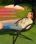 Woman relaxing on outdoor hanging lounge chair cushion with umbrella, curved hammock style, navy blue, patio or backyard setting