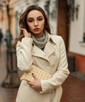 Woman in cream coat holding gold studded clutch, posing outdoors on a city street, styled for winter fashion.