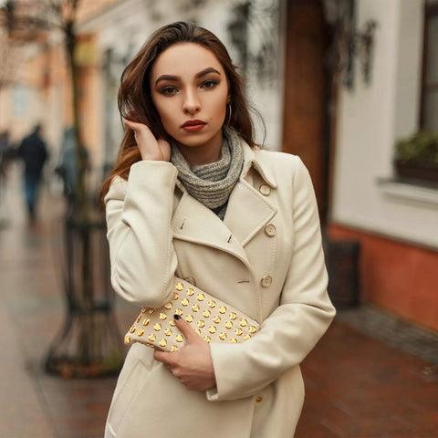 Woman in cream coat holding gold studded clutch, posing outdoors on a city street, styled for winter fashion.