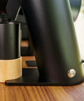 Solid Single Dose Coffee Grinder Featuring 63Mm Red Titanium Conical Burrs And Plasma Ionizer To Crush Beans Sick Flat And Eliminate Static Drama For Flawless Pulls In Sleek Black Housing, side view with wood stand.