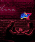 Two women sitting at a table under pink neon signs in a bar setting with brick wall backdrop.