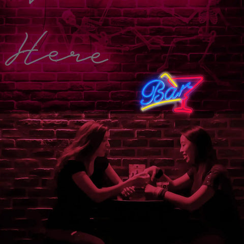 Two women sitting at a table under pink neon signs in a bar setting with brick wall backdrop.