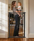 Woman holding a baby by the Extra Wide Adjustable Dog Gate For Doorways Built Tough Metal Pet Barrier in classic black.
