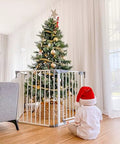 Child in Santa hat looks at Christmas tree behind Royale Converta 3 In 1 Play Yard And 151 Inch Wide Baby Gate, 6 panels.