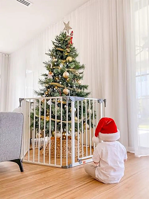 Child in Santa hat looks at Christmas tree behind Royale Converta 3 In 1 Play Yard And 151 Inch Wide Baby Gate, 6 panels.