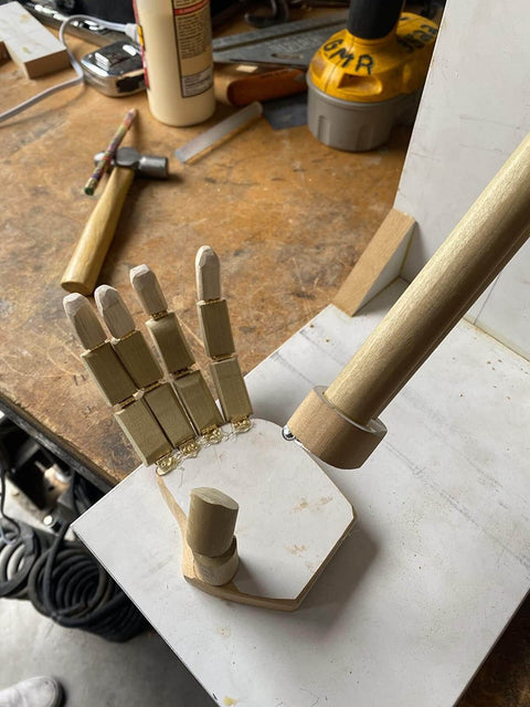 Wooden articulated hand model on a workbench with woodworking tools in the background, viewed from above.
