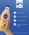 Game Changer Cocoa Butter Body Lotion For Crispy Dry Skin With Hyaluronic Acid And 72 Hour Deep Hydration To Melt Away Flakiness Reveal A Natural Glow And Leave Your Complexion Feeling Like Velvet All Day Long In A 16 9 Oz Tube held in a hand against a blue background.