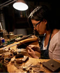 Person crafting jewelry using long needle nose pliers in a dimly lit workspace.