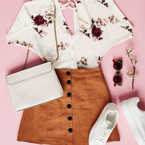 Flat lay of a floral blouse, tan skirt with black buttons, white sneakers, sunglasses, crossbody bag, and blossom sprigs on pink background