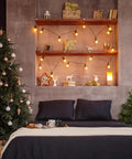 Cozy bedroom with Christmas tree, string lights on wooden shelves, and dark bedding against a textured wall.