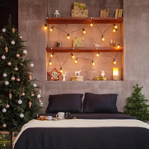 Cozy bedroom with Christmas tree, string lights on wooden shelves, and dark bedding against a textured wall.