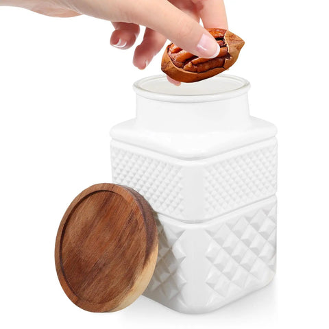 Hand placing nuts into a Vintage Embossed Matte White Glass Storage Canister with Rustic Wood Lid for kitchen organization.
