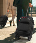 Black underseat roller backpack with wheels that pass airline size rules 18x14x8, water resistant, used by travelers on city sidewalk.