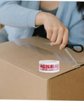 Person using Fragile Warning Packing Tape Set with 5 rolls to seal a cardboard box; heavy duty shipping tape for safe parcel handling.
