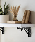Super Sturdy Matte Black Folding Shelf Brackets To Clear Your Clutter Fast These Compact Fold Down Wall Mount Supports Rock In Tight Spots Perfect For Garages Shops Or Kitchens Zero Sweat Two Pack, supporting a white shelf with decor.