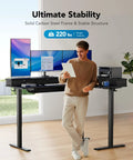 Man standing by 65 Inch L Shaped Standing Desk With Power Outlets And LED Strip, Electric Height Adjustable Corner Computer Desk for Home Office, Sit Stand Up Desk With Three Drawers, Black, holding tablet with monitors and printer on desk.