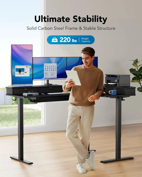 Man standing by 65 Inch L Shaped Standing Desk With Power Outlets And LED Strip, Electric Height Adjustable Corner Computer Desk for Home Office, Sit Stand Up Desk With Three Drawers, Black, holding tablet with monitors and printer on desk.