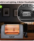 Gray SKOY laser engraver enclosure with built-in LED light and vent hose; front view with glowing orange interior.