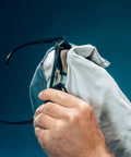 Glasses cleaning cloth, white microfiber, used to wipe black eyeglasses; close-up view.