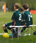 Two kids in soccer uniforms sitting on Six Foot Plastic Folding Bench Set With Handles, Portable HDPE Waterproof Outdoor Seating.