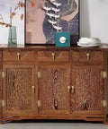 Wooden sideboard with intricate carvings and brass handles, placed under a decorative wall art, beside a vase and stacked plates.