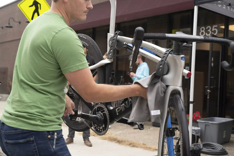 Man wiping a road bike on a repair stand with Waterless Ceramic Bike Cleaner Spray With Pina Colada Scent, 16 Oz, Quick Post Ride Shine, Safe On All Finishes, No Water Needed, Easy Touchup For Road, Mountain, And Gravel Bikes.
