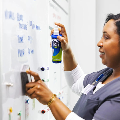 Woman using Killer Dry Erase Whiteboard Cleaning Spray on a whiteboard to remove stubborn ink and ghost marks.