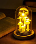 Mushroom Night Light In Glass Dome LED Decor With Wooden Base for cozy night ambiance on a wooden table next to open book.
