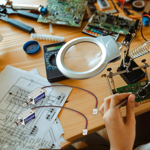 Two TEHAUX mini push-pull solenoids, 12V open-frame; side view with silver housings and blue ends on a wooden workbench.