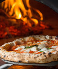 Pizza being removed from a wood-fired oven using a Thin Aluminum Pizza Peel With 13 And A Half Inch Wide By 16 Inch Long Surface And 8 Inch Wooden Handle For Indoor Oven And Outdoor Grillin Nonstick Transfer Paddle For Easy Pizza Bread And Pie.