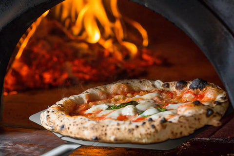 Pizza being removed from a wood-fired oven using a Thin Aluminum Pizza Peel With 13 And A Half Inch Wide By 16 Inch Long Surface And 8 Inch Wooden Handle For Indoor Oven And Outdoor Grillin Nonstick Transfer Paddle For Easy Pizza Bread And Pie.