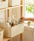 Person organizing beauty products in plastic storage bin with lid on wooden shelf by window
