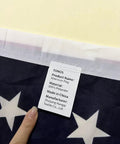 Close-up of the All American Patriotic 3x5 Ft Outdoor Flag With Brass Grommets label showing polyester material and manufacturer info.