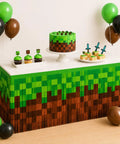 Two Pack Pixel Miner Party Table Skirts With Classic Green And Brown Block Pattern For Sick Gamer Birthday Decorations and Next Level 8 Bit Video Game Themed Party Supplies That Bring Ultimate Arcade Vibes To Your Party Table Setup, featuring a snack table with green and brown pixel design, cupcakes, and balloons.
