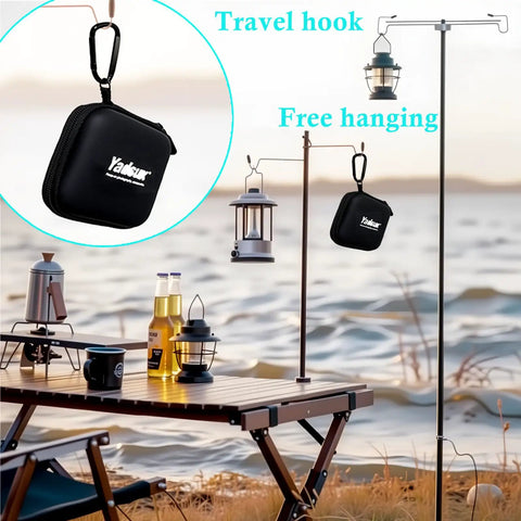 Outdoor scene with lanterns hanging from hooks and a black pouch with travel hook, near a lakeside picnic setup.