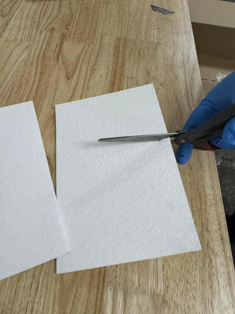 Two white ceramic fiber paper sheets being cut with scissors by a gloved hand.
