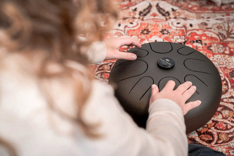 Person playing Super Responsive 16 Inch Steel Tongue Drum With Eight Soothing Notes Comes With Pro Grip Mallets And Carry Bag on a patterned rug.